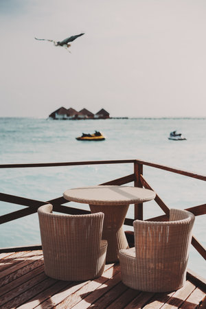 A vertical shot of a wicker table and chairs of a street bar of a luxury Maldives resort in the foreground and a bird, two water bikes, and a group of triangle bungalows in a defocused backgroundの写真素材