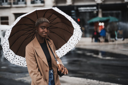 A portrait of a young cute black woman with short hair painted white, covering herself with a big patchy umbrella while standing on the rainy urban street, with a copy space area on the rightの写真素材