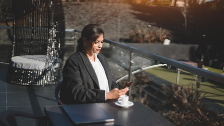 Side view of a beautiful plus-size woman using her cell phone while sitting in a street cafe during a coffee break with a cup of expresso and a closed laptop in front of her on the tableの写真素材