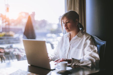 A portrait of a beautiful mature Hispanic female working with her laptop while sitting next to the window of a cafe; a plus-size cute Brazilian woman freelancer with netbook during her coffee breakの写真素材
