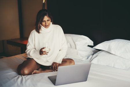 A portrait of a ravishing mature plus-size Hispanic woman in a white pullover watching a movie on her laptop while sitting on the bed of a cozy hotel room with a glass of white wine in her handの写真素材