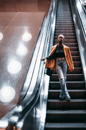 A vertical shot of a young elegant black female descending to an underground passage using a moving staircase; a cute black woman in a coat is going down on the escalator to the subway station depotの写真素材