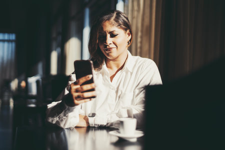 Portrait of a charming plus-size Hispanic business lady sitting in a dark interior of a restaurant and replying in a chat using her smartphone; a cup of espresso coffee next to her on the tableの写真素材