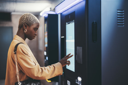 A side view of a young beautiful elegant black woman with painted white very short hair paying for service underground parking or buying a subway or train ticket using an electronic self-service kioskの写真素材