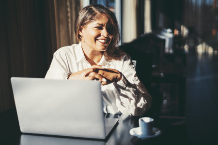 A portrait of a cheerful mature Hispanic plus-size woman entrepreneur in a white shirt sitting at a cafe table with her laptop and a cup of espresso during her coffee break, smiling and looking asideの写真素材