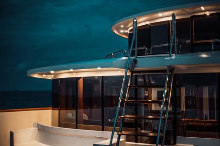A night low-key shot of a luxury diving safari yacht deck lit by multiple ceiling lamps, with a ladder in front and a glass row of windows with a wardroom behind it; a twilight sky in the backgroundの写真素材