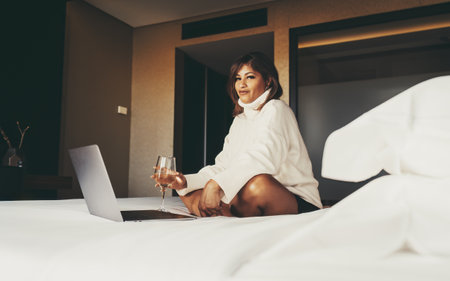 A mature beautiful plus-size Hispanic female in a pullover is sitting on the bed in the interior of her cozy bedroom with a glass of white wine and surfing the net via a laptop in front of herの写真素材