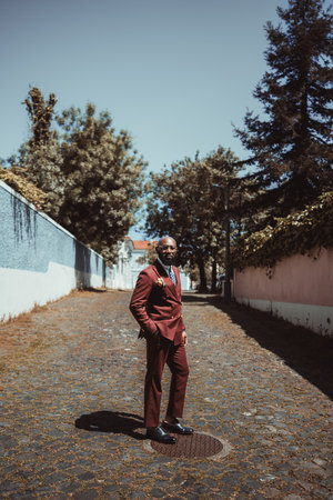 A vertical shot of an elegant mature bald African senior with a well-groomed black beard, in eyeglasses and a chestnut color, tailored suit with a necktie, standing on the paving stone on the streetの写真素材