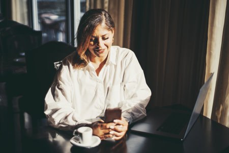 A portrait of a plus-size Caucasian woman entrepreneur is sitting in the dark interior of a restaurant lit by contrast sunlight from the window, using her smartphone and a laptop; a coffee breakの写真素材