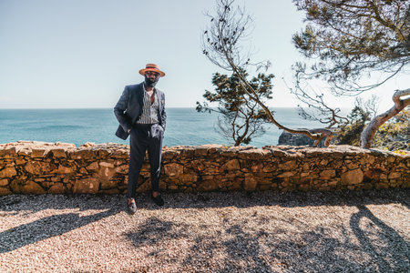A wide-angle view of an elegant bearded mature black man in a straw hat and a tailored summer costume of a retro style with a striped shirt, standing on the shore next to the ocean and a stone fenceの写真素材