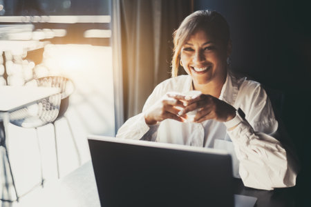 Portrait of a happy smiling Hispanic female entrepreneur in a white shirt, sitting with a cup of expresso coffee in her hands in front of her laptop next to the window of a cafe or a restaurantの写真素材