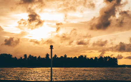 View from the oceanÂ and a vessel on a dark silhouette of a tropical island shoreline overgrown with palm trees, with and stunning skyscape above during a dramatic sunset and golden hour; Maldivesの写真素材