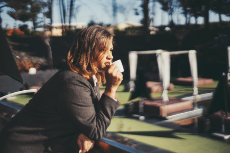 A side view of a Hispanic woman drinking espresso while leaning against fencing; profile view of a plus-size Caucasian female with golden hair drinking coffee outdoors; a copy space area on the rightの写真素材