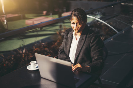 Portrait of a ravishing adult plus-size caucasian woman entrepreneur working on her laptop while sitting in a street cafe with a cup of espresso coffee on the table next to her on a windy autumn dayの写真素材
