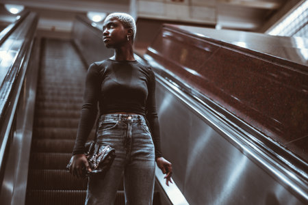 A low-key portrait of a young cute black female with very short painted white hair holding a clutch bag while standing on the escalator and looking aside; an African woman on the moving staircaseの写真素材