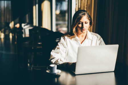 A contrast portrait of a dazzling mature plus-size Hispanic businesswoman in a white shirt sitting indoors at a restaurant and using her laptop with a cup of expresso coffee next to her during a breakの写真素材
