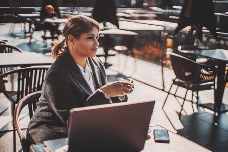A side view of a mature plus-size Hispanic woman entrepreneur with a laptop and a cup of coffee in her hands, sitting at the table of a street restaurant and having a break during a freelance projectの写真素材