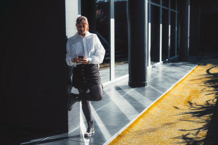 A plus-size Hispanic businesswoman leaning against the black wall of an office building exterior with a strict glass facade and using her smartphone, with a copy space area on the rightの写真素材