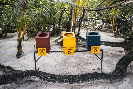 Three trash containers for separate waste collection raised above the coral sand in tropical settings: a red one for metal, yellow for plastic, and blue for paper, with a tree trunk in frontの写真素材
