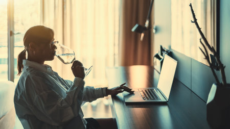 A low-key side view of a mature plus-size Hispanic female drinking wine and using her laptop while sitting and having a rest in a dark hotel room during her vacation tripの写真素材