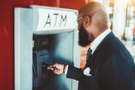 A side view of an elegant bearded bald black man entrepreneur in a suit and glasses with a selective focus on his hand inserting a credit card into an outdoor bank ATM to withdraw some cashの写真素材