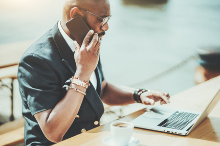 A side view of a serious black businessman talking on the phone while sitting with his netbook in a street cafe; an African man freelancer on a patio using his laptop during a coffee breakの写真素材