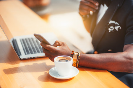 Close-up view of the hand of a black man entrepreneur in a formal summer costume using a smartphone in a street cafe, with a cup of coffee and a laptop next to him on the table on a warm sunny dayの写真素材