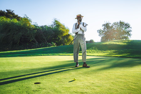 View of a fashionable handsome bearded black senior with a sigar, in straw hat and tailored outfit standing on a green golf field with a club in his hand; a yellow golf ball and a hole in front of himの写真素材