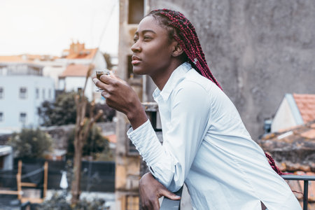A side profile view of a charming youthful African female with long red box braids leaning against the fence of a cafe balcony or her house fencing while drinking a delicious expresso coffeeの写真素材