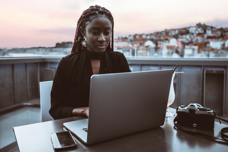 A portrait of a dazzling young black femaleÂ hipster freelancer with red box braids working on her laptop while sitting on the roof cafe with a cityscape and pink sunset in a defocused backgroundの写真素材