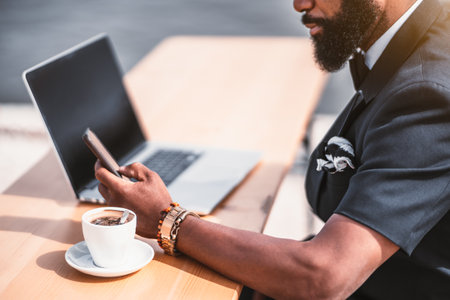 A bearded black African businessman in a street cafe, with a selective focus on his hand typing a message on the cellphone; a cup of espresso coffee next to him on the wooden table near his netbookの写真素材