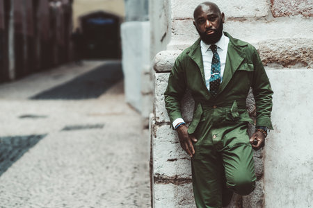 A dapper tough bald bearded handsome black guy in a fashionable greenish outfit leaning against the wall outdoors; a street portrait of an African man in elegant green overalls with a necktieの写真素材