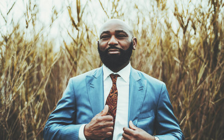 Portrait of a fashionable bald mature black man entrepreneur with a thick beard and in an elegant blue suit with a necktie is adjusting the blazer while standing outdoors with reeds in the backgroundの写真素材