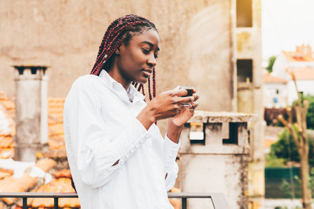 A charming young African American female with dark-red braided hair and in a white shirt is drinking an espresso coffee while standing on the balcony of her apartment lit by a warm morning sunの写真素材