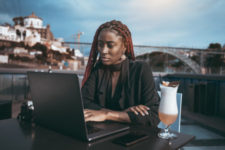 A portrait of an occupied ravishing black businesswoman using her laptop while sitting in a street cafe with a cocktail; a dazzling African American woman entrepreneur outdoors with a netbookの写真素材