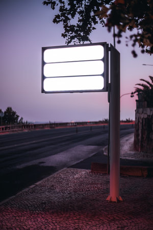 A vertical selective focus capture of three empty white color street sign templates by the roadside of the sidewalk with the purple dusk sunset in the backgroundの写真素材