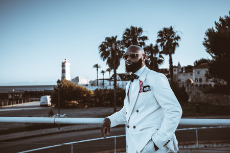 An irresistible bald, bearded black businessman, in a fashionable suit, sunglasses, and a polka dot bow tie standing against a metal fence on a terrace with palm trees and a lighthouse in the backの写真素材