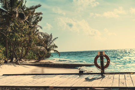 View of the end of a wooden boardwalk with a little bit of sand on it, pinned to the path a red ring buoy; surrounding it teal ocean water, inclined trees, and waves on a somewhat cloudy but warm dayの写真素材