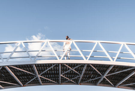 Low-angle shot of a dandy bald African American executive dressed mono-chromatically in a white suit with a well-groomed beard has his arm resting on the white metal bridge fencing on a bright dayの写真素材