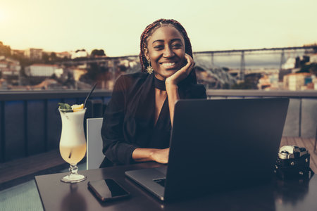 A portrait of a young cheerful African American woman entrepreneur using her laptop while sitting in an outdoor restaurant; a dazzling smiling black female freelancer in a street cafe with a netbookの写真素材
