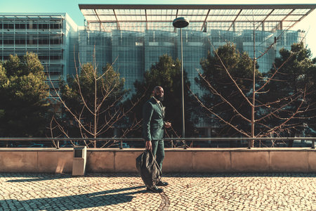 A sophisticated Angolan male was captured outside of the office building, in the back, standing on the pavement wearing a chequered dark green suit and carrying a weekender on a sunny dayの写真素材