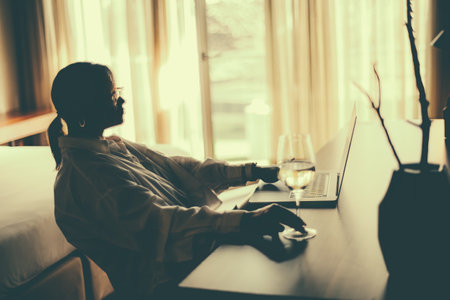 A silhouette of a curvy woman sitting at the table with her laptop and a wineglass with white wine and watching a film in a dark hotel room, with a bed and a balcony in the background; selective focusの写真素材