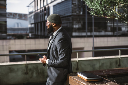 A profile shot of a middle-aged black man with an unshaven beard, eyeglasses, and a dark cap, holding a smartphone and leaning against a vase with an olive tree and a notebook sitting on top of itの写真素材