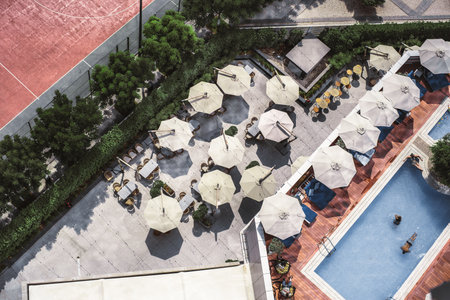 Top view from a drone of an outdoor bar with sunshades creating a beautiful landscape, adjacent to a lounge and pool area with people resting, bordering the terrace trees hide the clay tennis courtの写真素材