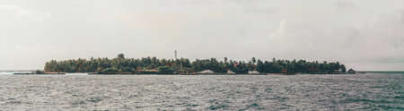 A tropical island landscape in the Maldives is captured in a panoramic view, featuring lush vegetation, calm blue seawater, and an overcast gray sky, creating a serene and warm atmosphereの写真素材