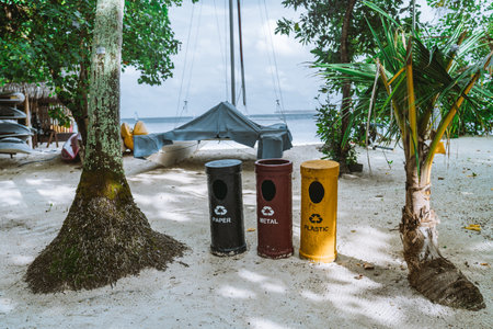 Three eco-friendly cylindrical recycling bins, colored in yellow, red, and blue for plastic, metal, and paper respectively, were placed on a beach surrounded by tropical trees with varied foliageの写真素材