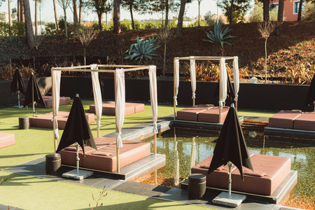 An idyllic natural pool with outdoor loungers draped in warm earthy hues, seamlessly blending with the lush surroundings of trees and ground, while a closed black beach umbrella provides shadeの写真素材