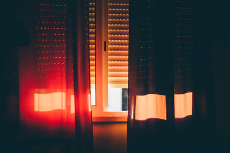 In the afternoon, the bedroom is drenched in a warm orange glow as sunlight seeps in through the partly drawn blinds and filters through the red curtains fluttering in the breeze from the open window.の写真素材