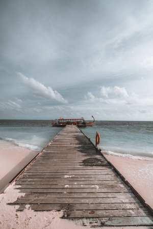 A vertical shot of a very long wooden pier on a Maldivian beach island, leading to a simple yet beautiful gondola boat moored in the gray seawater on a day with clouds in the skyの写真素材