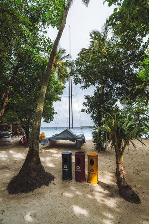 A vertical wide-angle shot in the Maldives with eco-friendly trash bins on a sandy beach surrounded by palm trees, and the boat in the background promotes sustainable tourism and natural beauty.の写真素材